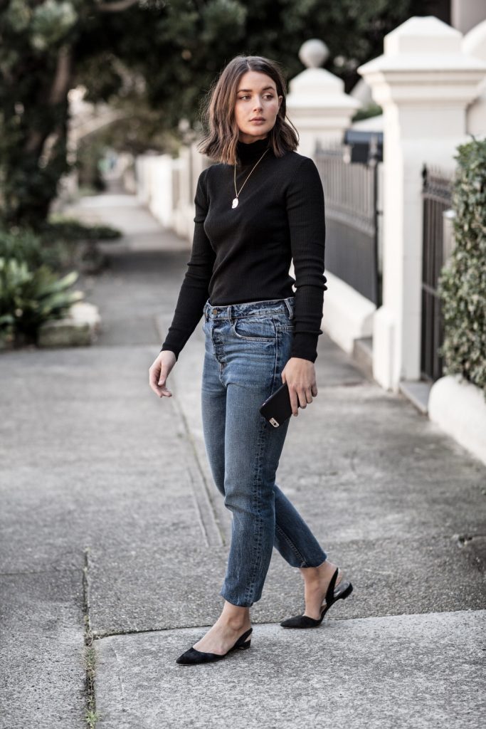 Woman standing on footpath wearing black turtleneck and denim jeans