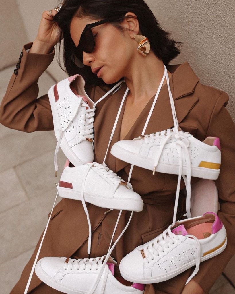 Woman in sunglasses wearing white Bared Footwear sneakers around her neck