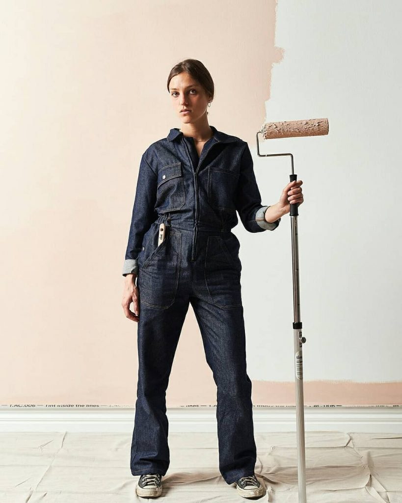 woman in overalls standing with a large paint roller in front of a half painted wall mid-home renovationslf 